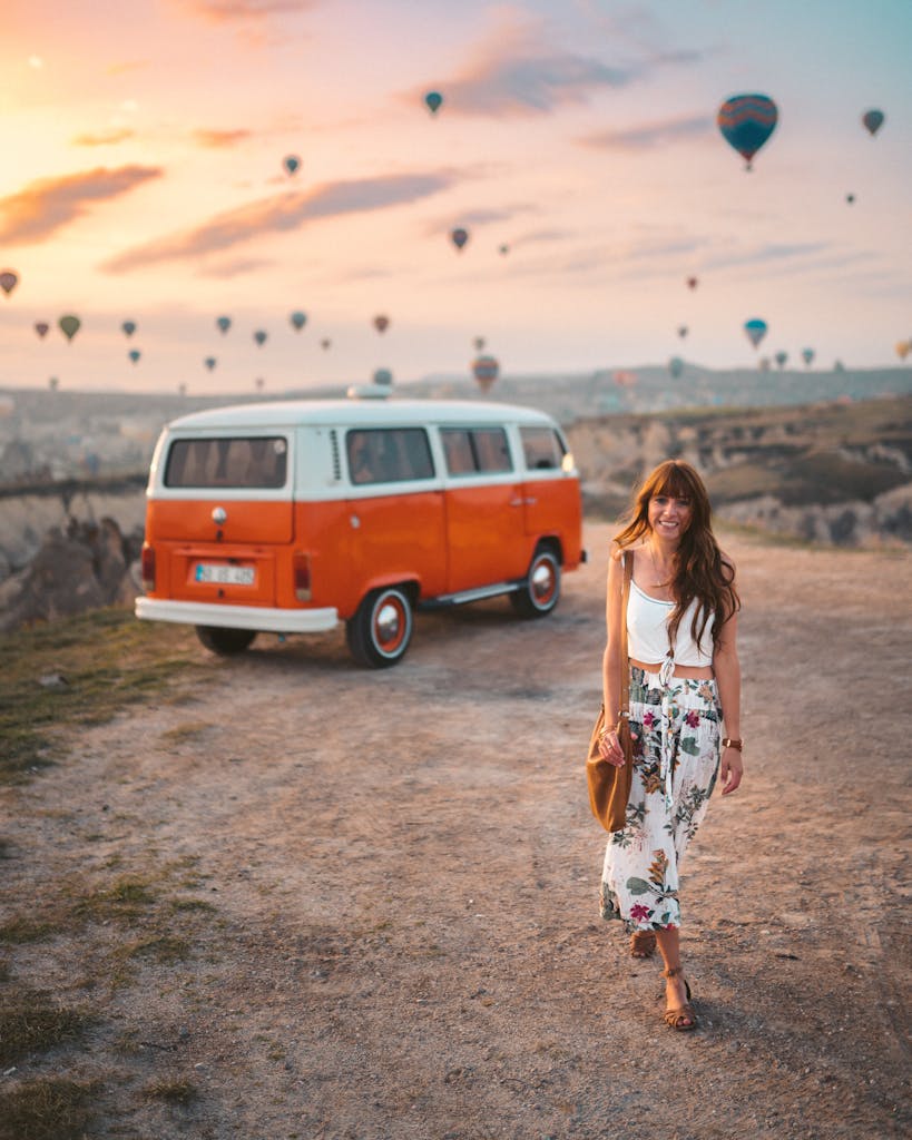 A woman walking by an orange van during a vibrant sunset in Cappadocia with hot air balloons in the background.