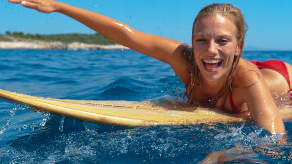 CLOSE UP, PORTRAIT, DOF: Smiling surfer girl having fun swimming to the line up to catch big crashing waves. Beautiful young Caucasian woman enjoys her summer vacation by surfing on a perfect day.
