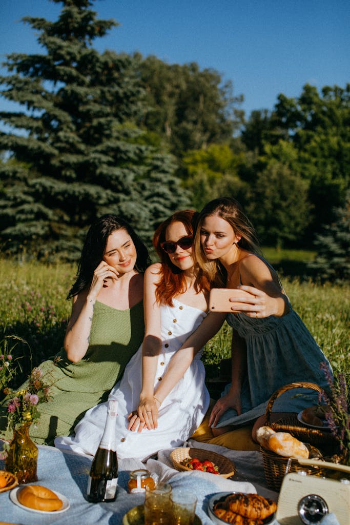 Three women enjoying a summer picnic in a park while taking a selfie together.