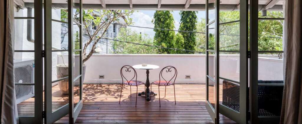 Green-framed French doors open onto a private timber-decked roof terrace with a marble bistro table, two red heart-backed iron chairs and a frangipani tree, looking out over Galle Fort's treetops